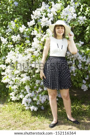 European lady in white hat standing near big lilac shrub in blossom.