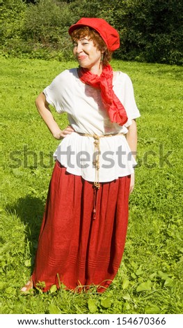 European woman in red hat, white blouse and red skirt  with red scarf standing on grass meadow in park, forest behind.