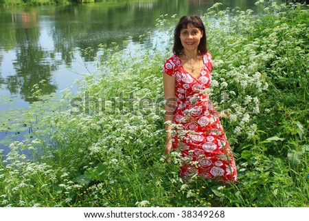 Girl (young woman), black hair, red dress, standing on bank of lake, piece of water, white wild flowers around.