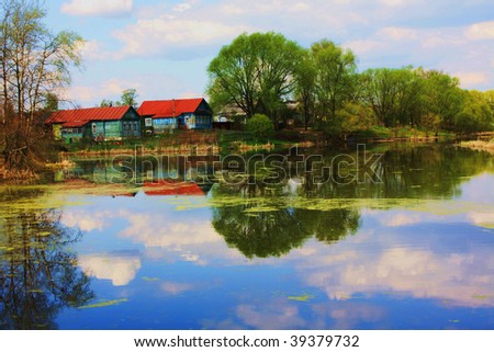 Summer landscape - wooden houses on bank of lake, trees, reflection of houses, plants and clouds in water.