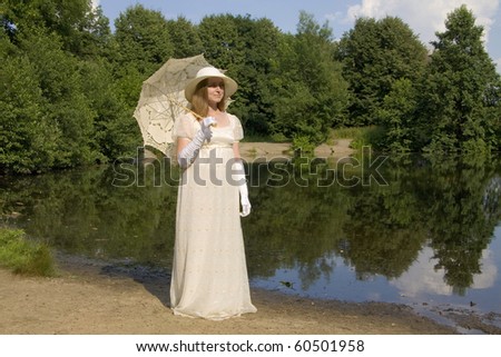 Young woman in white historical dress of XIX century with white umbrella in white hat near lake.