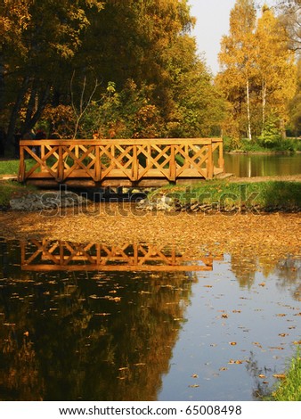 Autumn landscape with bridge on the lake, little island with birch trees, forest around, reflection in water. Recorded in Sokolniki park in Moscow.