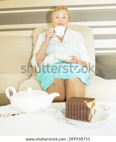 European blond lady in ages drinking tea at home, cup of tea in hands, catle and biscuit on table.