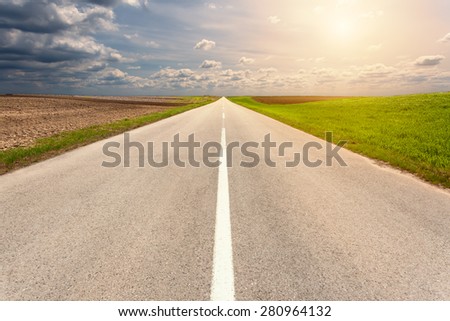 Driving on an empty asphalt road through the cultivated fields towards the setting sun