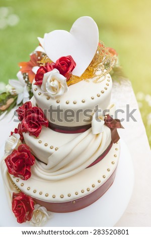 Elegant wedding cake on three floors with edible decoration in the shape of roses, ribbons and heart in nature environment, illuminated by the sunlight.