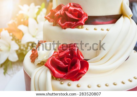 Closeup of elegant wedding cake with edible decoration in the shape of roses and bouquet of flowers in background, illuminated by the sunlight.