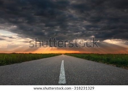 Driving on an empty asphalt road through the countryside fields towards the setting sun and sunbeams.