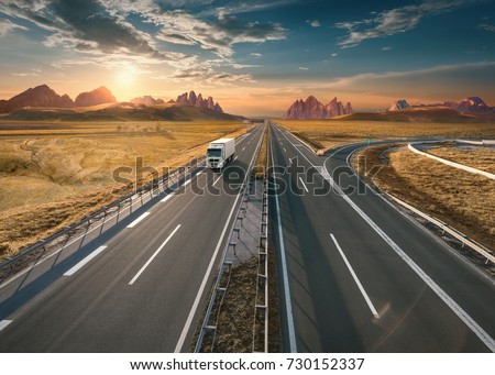Single delivery truck driving from the mountain range towards the setting sun. Fast motion drive on the straight freeway in beautiful landscape. Freight scene on the empty motorway.