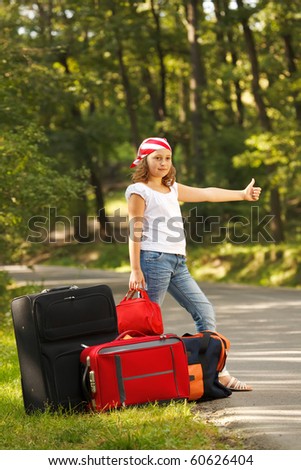 Young hitch-hiker girl standing on road side afternoon in forest with bags