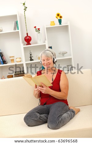 Happy senior woman sitting on sofa, reading book and listening to music