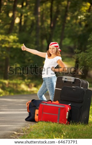 Young hitch-hiker girl standing on road side afternoon in forest with bags