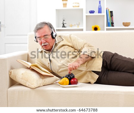 Senior man laying on sofa, reading and listening to music while eating fruits