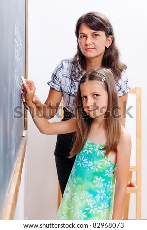 Teacher standing behind elementary, helping her to write on chalkboard by driving her hand