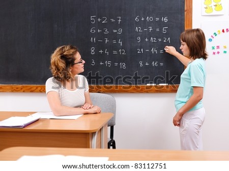 Elementary student solving equations at chalkboard while teacher looks at her