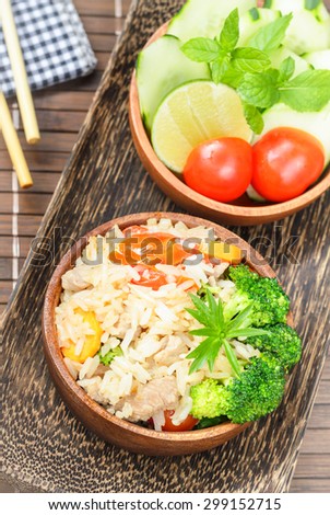 Healthy eating, homemade stir fried rice with vegetable, meat and egg served on wooden bowl and side bowl of fresh cucumbers, tomatoes on wooden plate with chopsticks (Selective Focus, Focus on rice).
