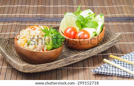 Healthy eating, homemade stir fried rice with vegetable, meat and egg served on wooden bowl and side bowl of fresh cucumbers, tomatoes on wooden plate with chopsticks (Selective Focus, Focus on rice).