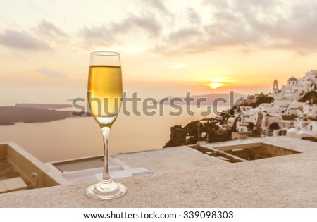 a glass of white wine on balcony with Caldera city view at sunset, yellow sunlight and bright sky before getting dark (selective focus on a glass of wine)