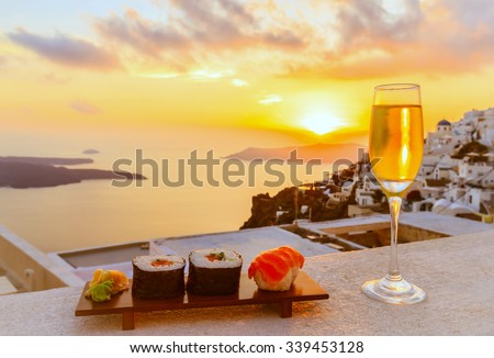 Sushi and a glass of white wine on balcony with Caldera city view at sunset, yellow sunlight and bright sky before getting dark (selective focus on sushi)