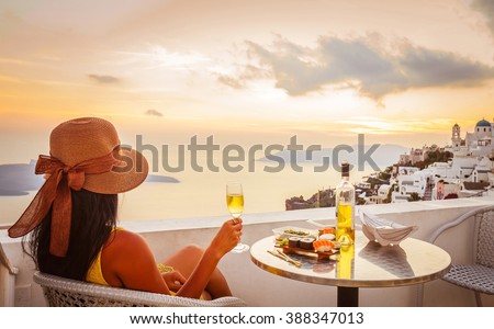 Young woman wearing hat, looking at beautiful view of Caldera and enjoying romantic dinner during sunset before getting dark over Aegean sea, Santorini