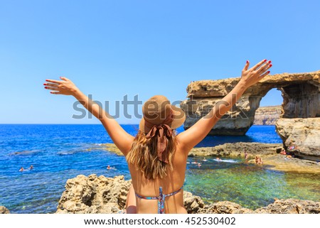 Back of female tourist wears hat, bikini, raises hands up and enjoys summertime with background of Azure window, famous tourist attraction on Gozo island, Mediterranean sea, Malta