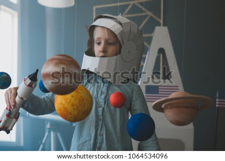 Cute little girl wearing cardboard astronaut helmet flying toy rocket through planets, cardboard spaceship rocket in the background