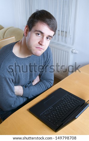 high angle view of upper body of a young man with laptop and arms folded on the table sitting and surly looking into the camera