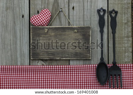 Blank wood sign by gingham tablecloth and cast iron spoon and fork hanging on wood background
