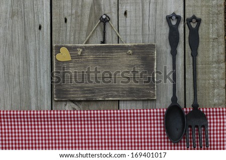 Blank wood sign by gingham tablecloth and cast iron spoon and fork hanging on wood background