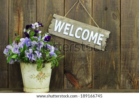 Welcome sign with potted flowers (purple pansies) by wooden fence