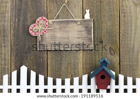 Blank wood sign with plaid heart over white picket fence and birdhouse