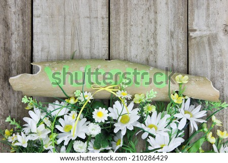 Green weathered welcome sign with border of flowers and daisies by wooden fence