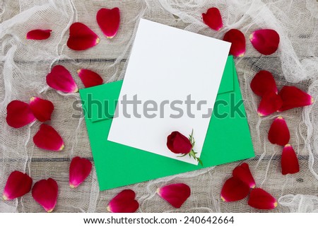 Blank white letter or note card and green envelope with red flower and red rose petals on shabby white netting background