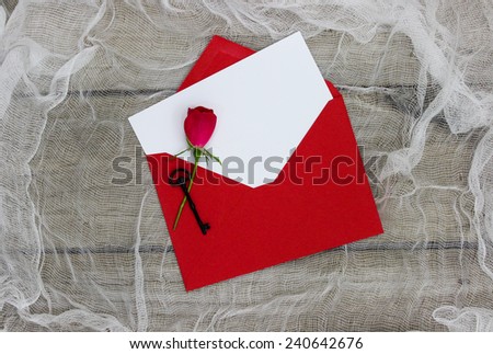 Blank white love letter or note card with red envelope, red rose and black iron key on shabby white netting and wooden background