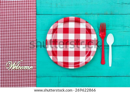 Welcome sign by red and white checkered picnic plate with gingham tablecloth on antique teal blue wooden table; above view looking down