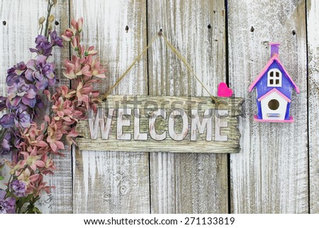 Welcome sign hanging on white fence by spring flowers and purple and pink birdhouse