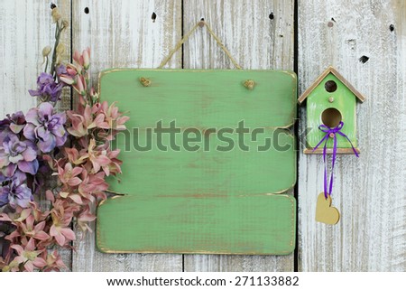 Blank wood green sign hanging on white fence by spring flowers and green birdhouse with hearts