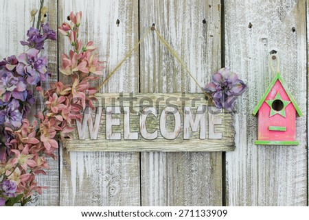 Welcome sign hanging on white fence by spring flowers and pink birdhouse