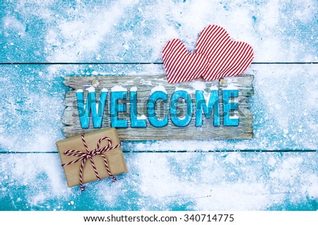 Holiday welcome sign with present and candy cane striped hearts on teal blue snowy wood background