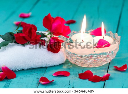 Spa composition with red flowers and rose petals, white towel, aroma bowl with three white floating candles on antique rustic teal blue wood background