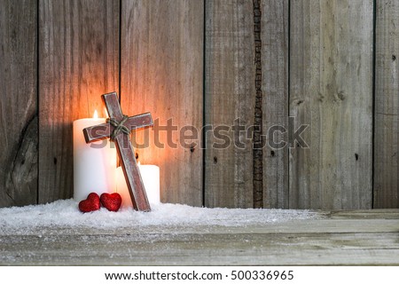 White holiday candles glowing behind wooden cross with red hearts in snow by antique rustic wood background; Christmas and religious background with copy space