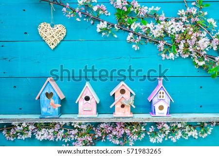Natural wicker heart hanging over colorful birdhouses with butterfly on shelf by spring tree flowers on antique rustic teal blue wooden background; springtime background with painted wood copy space