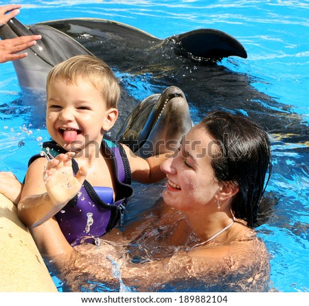 Happy beautiful young woman with a small child laughs and swims with dolphins in blue swimming pool on a clear sunny day