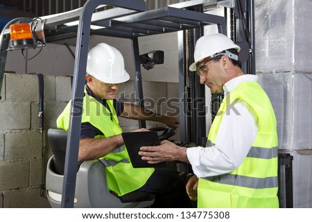 Two men wearing safety gear working in a warehouse. One man showing the other something on a digital tablet.