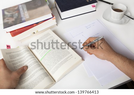 A male student is reading a book and taking notes. Coffee and books on the desk