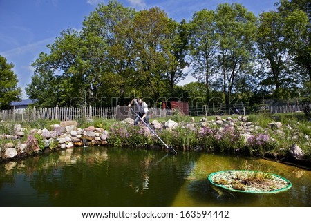man fishes in garden pond with a landing net, a stone waterfall and pasture fences in the background