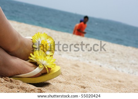 Yellow flowered sandals relax on a sandy beach, with the ocean in the background.  It\'s sunny, hot and relaxing.