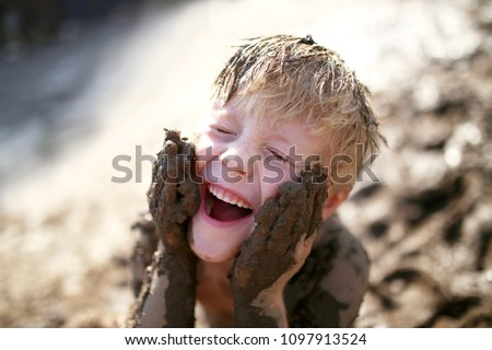 A cute little boy child is laughing as he plays outside in the mud and rubs dirt on his face with his hands.
