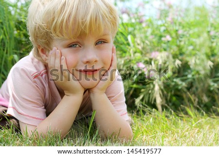 A cute little boy child lays outside in the grass by some flowers on a summer day, resting his head in his hands and smiling a small smile at the camera