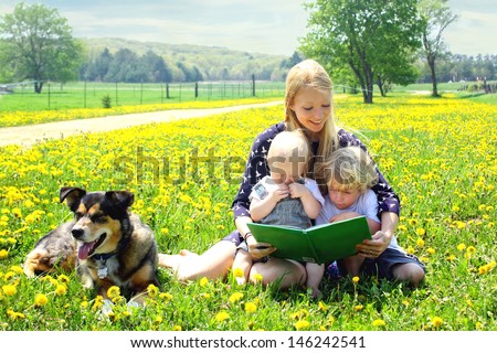 a young attractive mother reads a story book to her two small children while sitting outside in a meadow with their German Shepherd dog