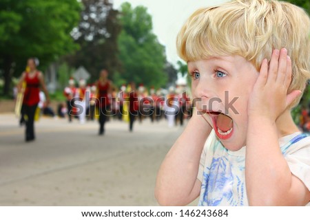 A cute toddler boy covers his ears as he watches a school marching band walk by in a parade on a summer day